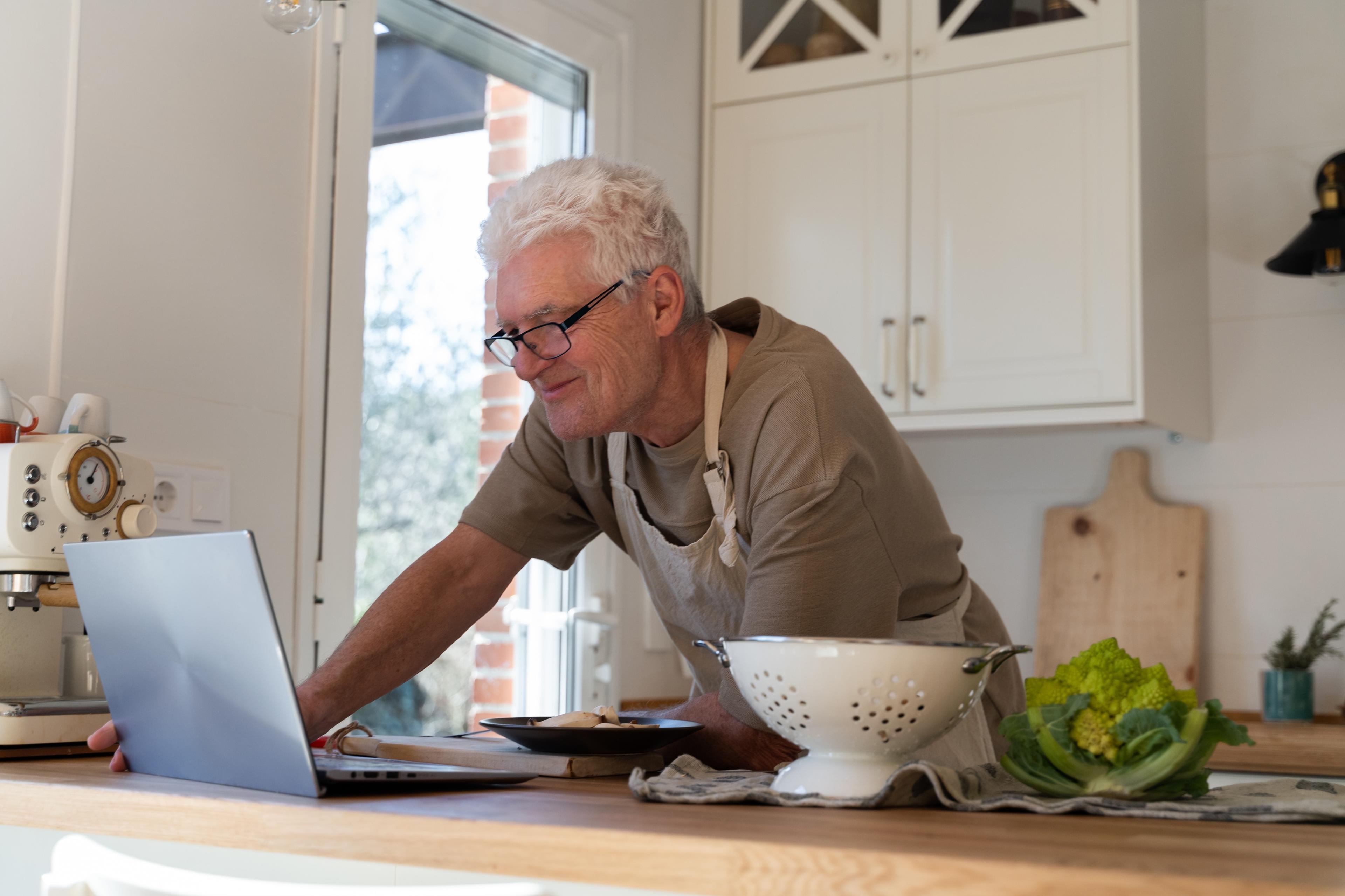 A middle-aged man wearing an apron stands at his kitchen counter surrounded by a variety of healthy ingredients. He smiles while looking at his laptop during a virtual call with his dietitian.